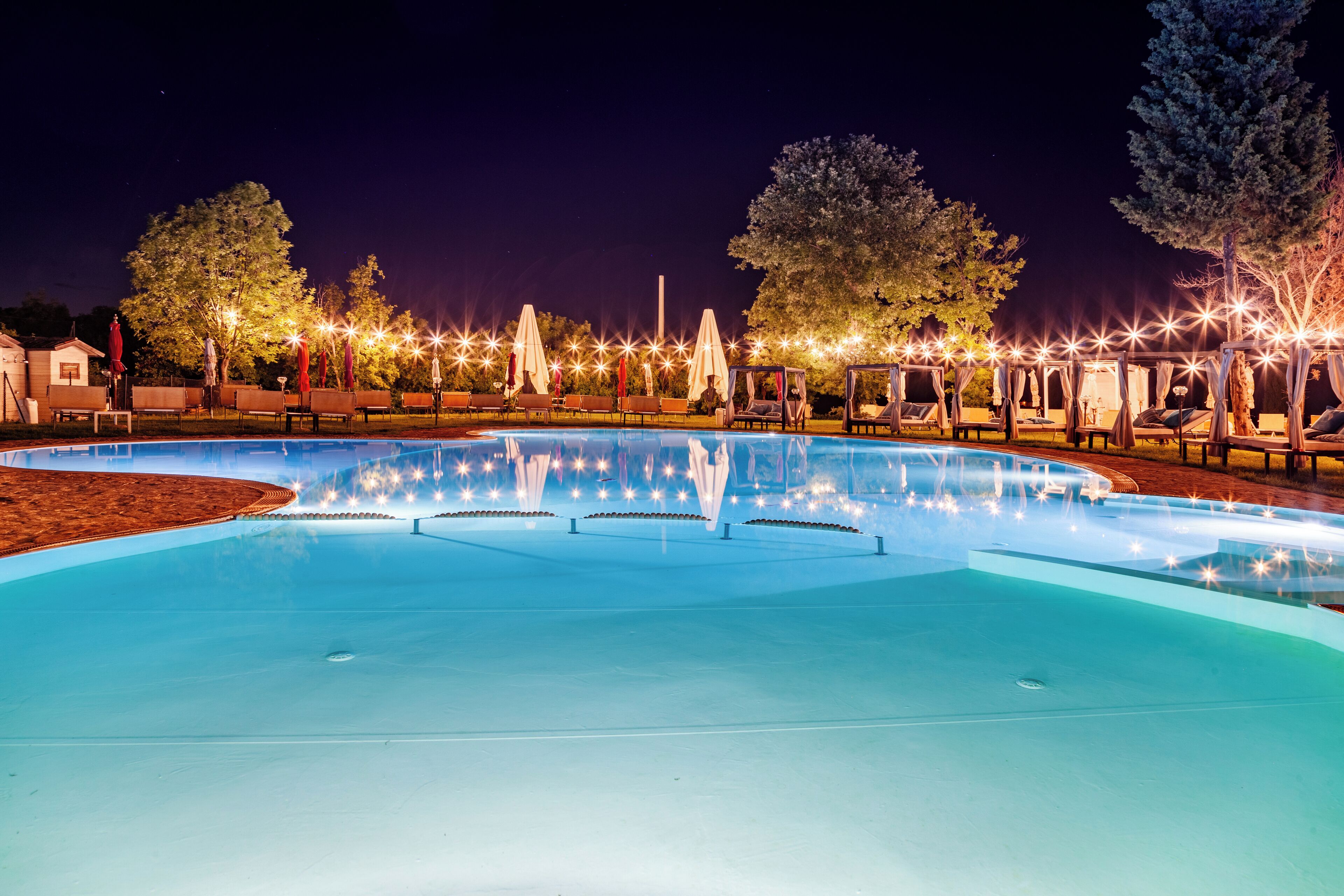 Una piscina con sillas de descanso y sombrillas, rodeada de árboles e iluminada con luces colgantes.