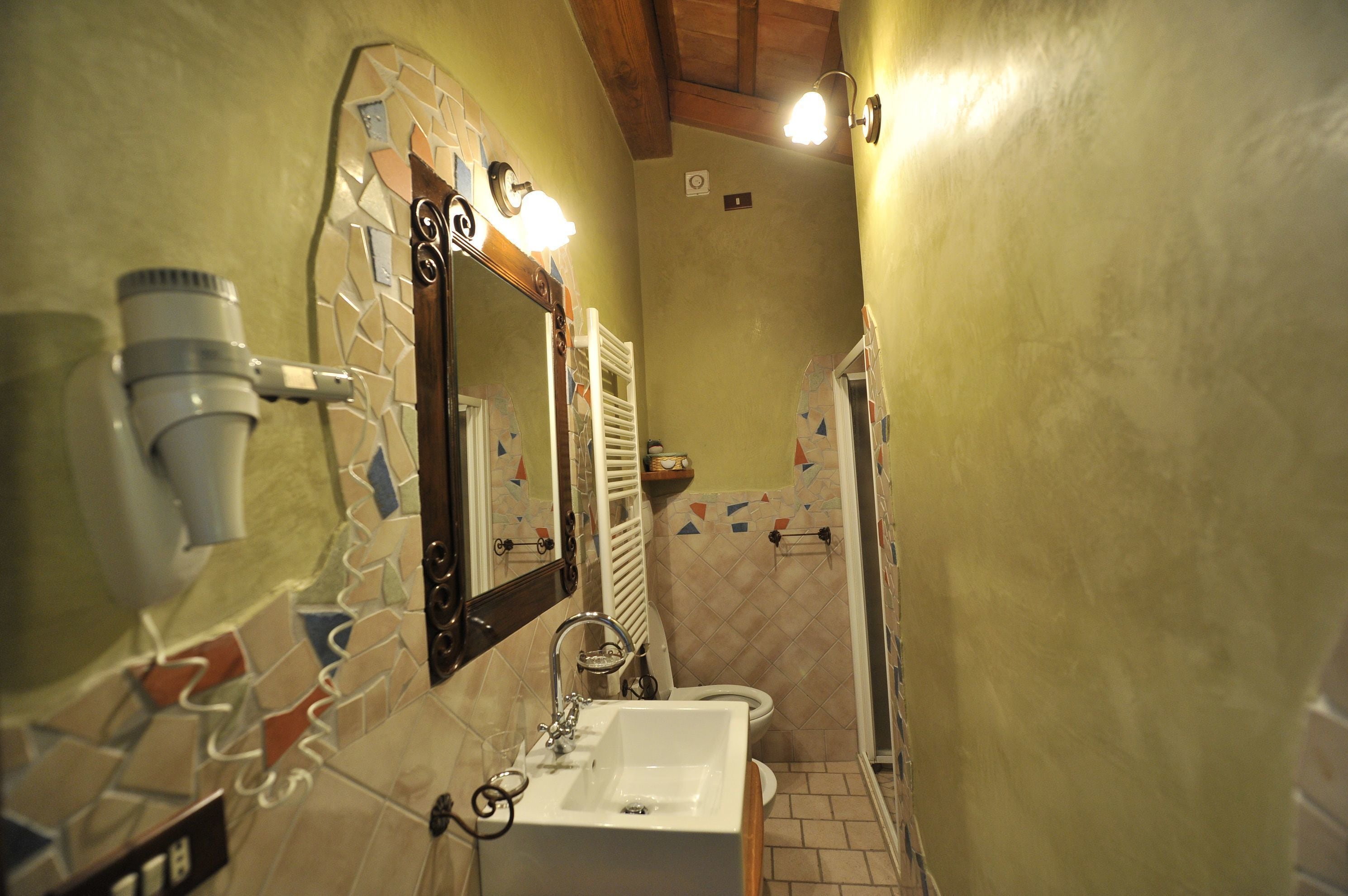 Baño con lavamanos blanco, espejo y toallero. Hay un secador de pelo en la pared y una zona de ducha con pared de azulejos.