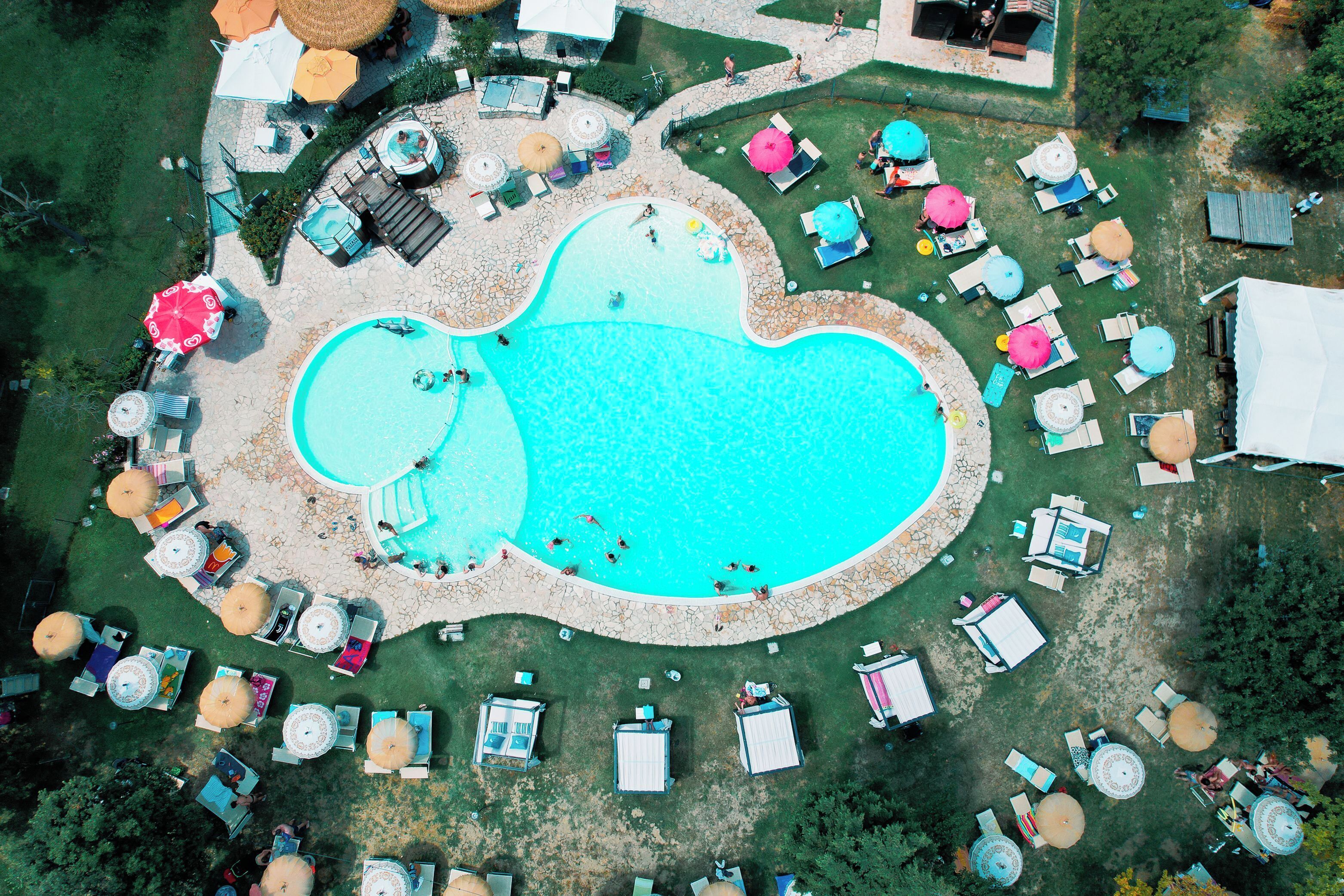 An aerial view of a resort with a large central pool, multiple lounge areas with umbrellas, and a well-maintained lawn.