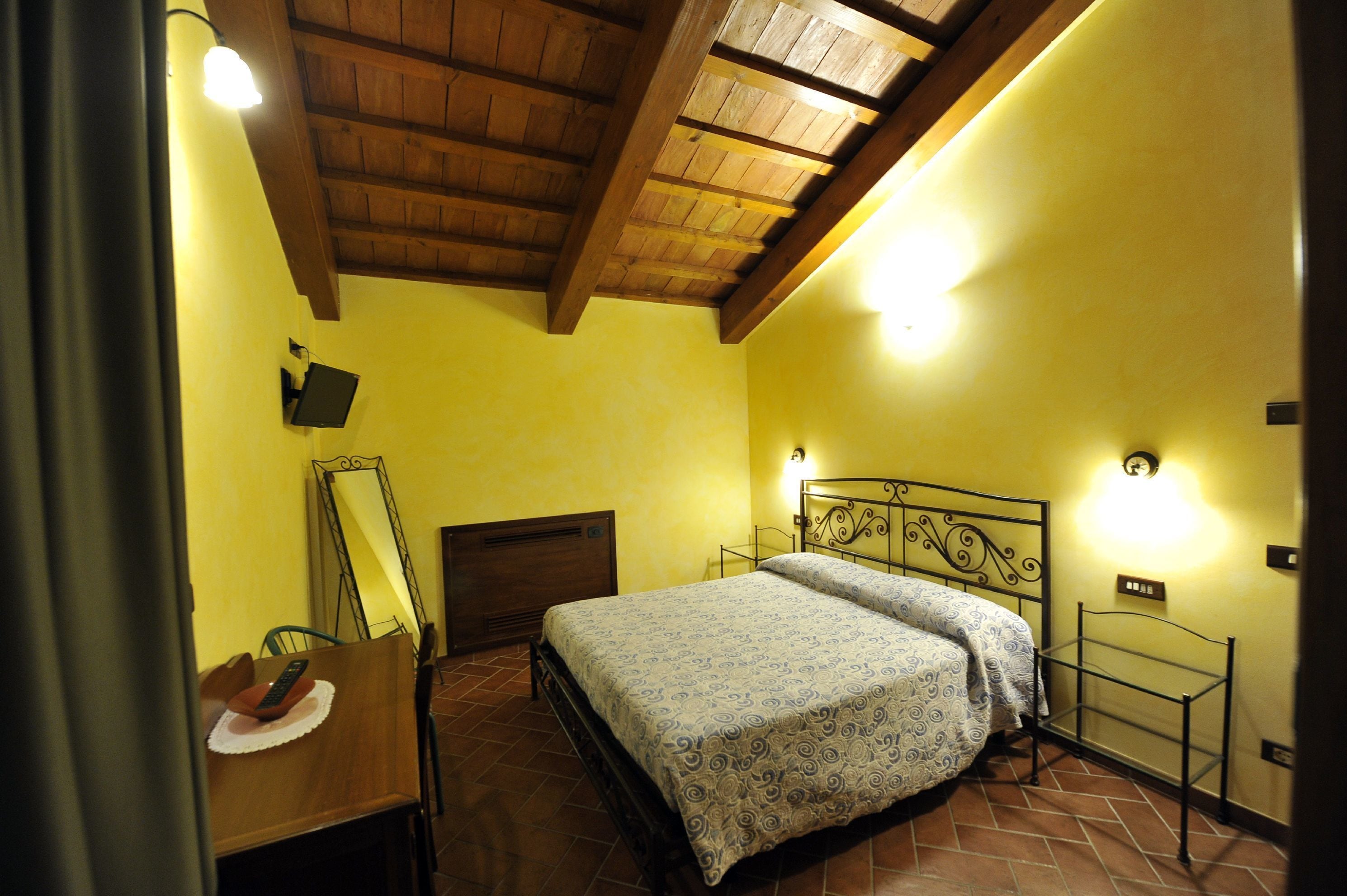 A bedroom with a wooden ceiling, a bed with patterned bedding, a bedside table with a lamp, and a wall-mounted mirror.
