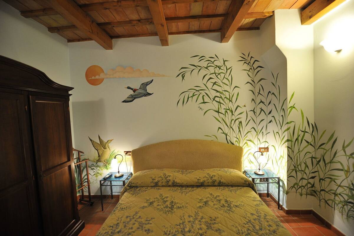 A bedroom with a bed, a wooden wardrobe, a wall mural, and a nightstand with a lamp.