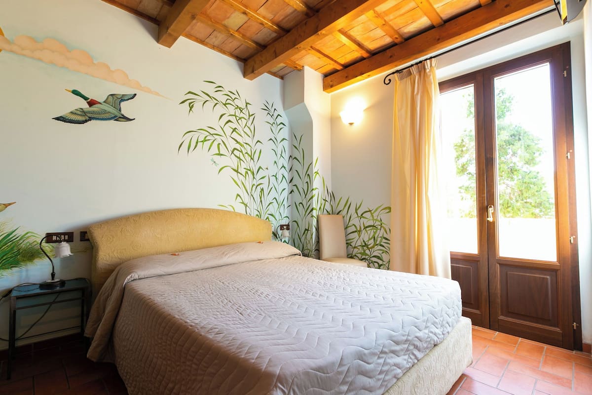 A bedroom with a bed, a nightstand, a wall-mounted lamp, a large window with curtains, and a wooden ceiling.