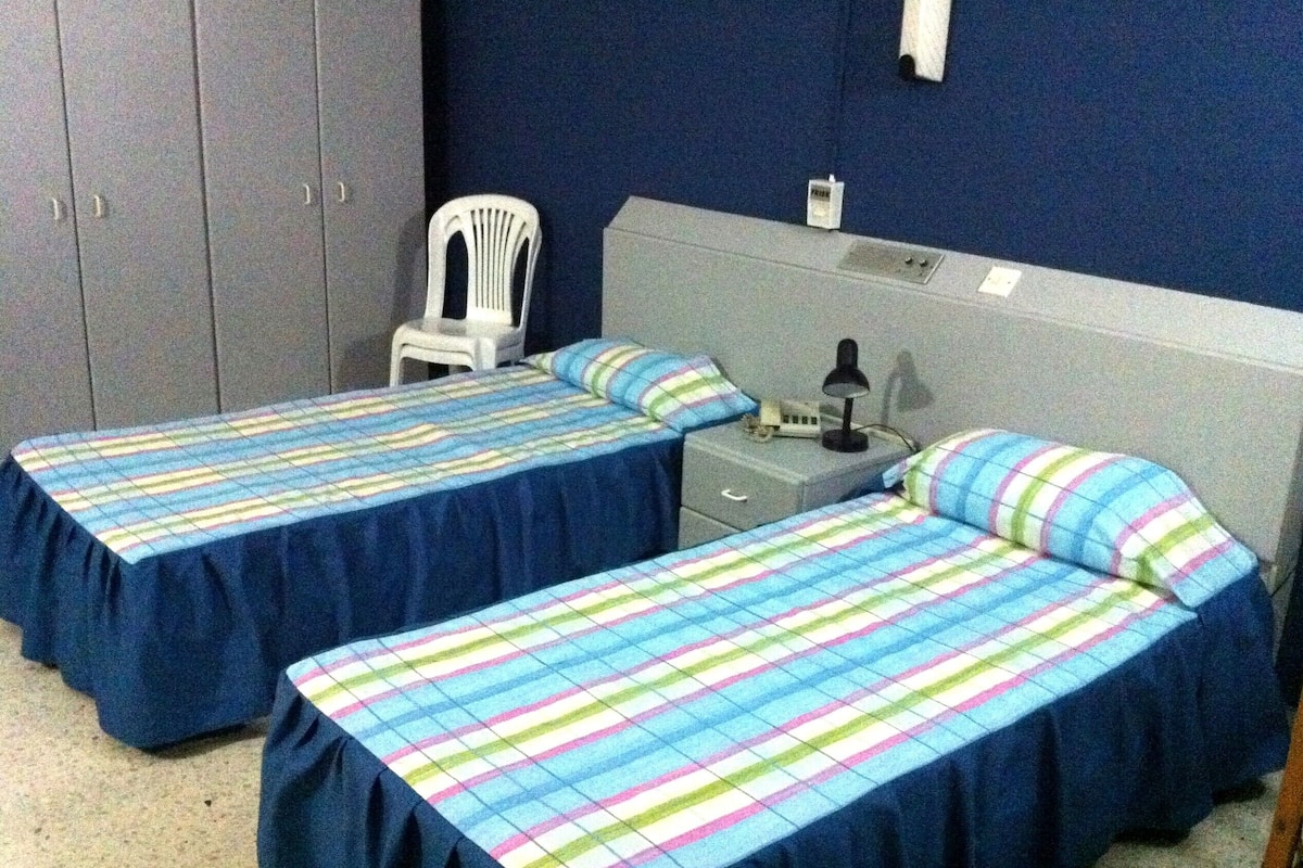 A room with two single beds, a nightstand with a lamp, a chair, and a wardrobe.