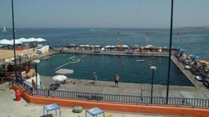 A waterfront promenade with a pool, umbrellas, and people. - Hotel Roma (Sliema)