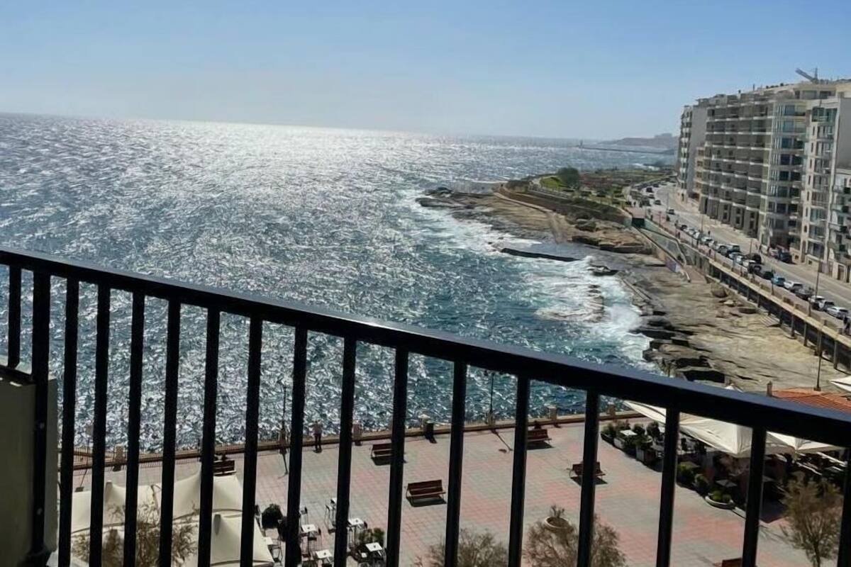 A coastal view from a balcony with a railing, overlooking a rocky shoreline and a cityscape with buildings and parked cars.