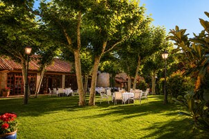 Un área de comedor al aire libre con mesas y sillas, rodeada de árboles y un edificio con techo de tejas.
