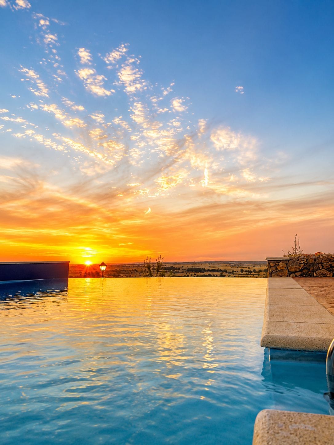 Vista del atardecer sobre una piscina con un muro de piedra y farol.