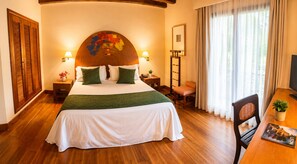 Habitación de hotel con una cama grande, piso de madera, un escritorio con silla, televisor y ventana con cortinas.