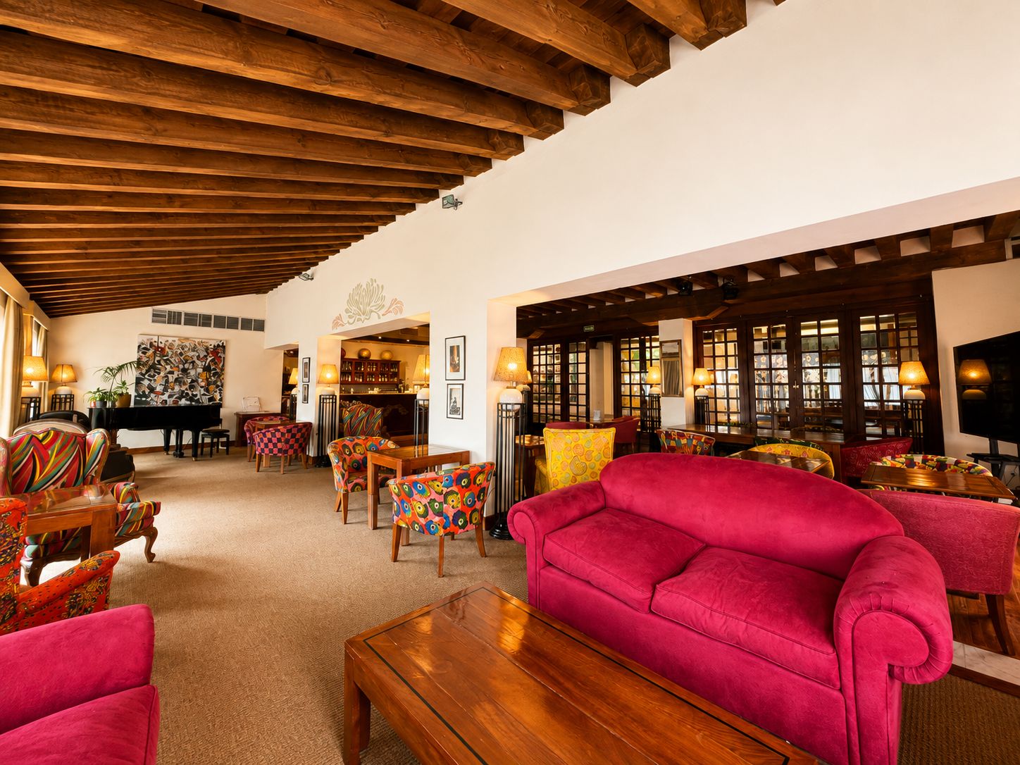 Un vestíbulo de hotel con techo de madera, un piano, sillas de colores, un sofá rojo y una zona de bar.
