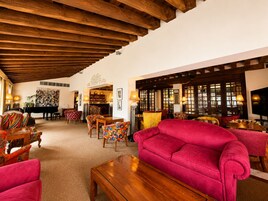 Un vestíbulo de hotel con techo de madera, un piano, sillas de colores, un sofá rojo y una zona de bar.
