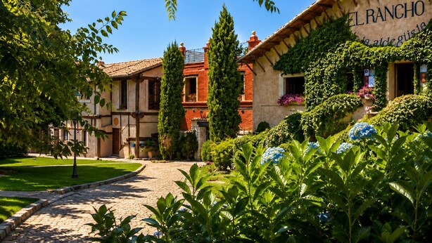 Un patio con un camino adoquinado, un edificio con la leyenda 'EL RANCHO' y abundante follaje.