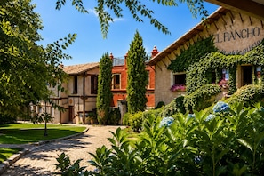A courtyard with a cobblestone path, a building labeled 'EL RANCHO,' and lush greenery. - Hotel El Rancho (Torrecaballeros)