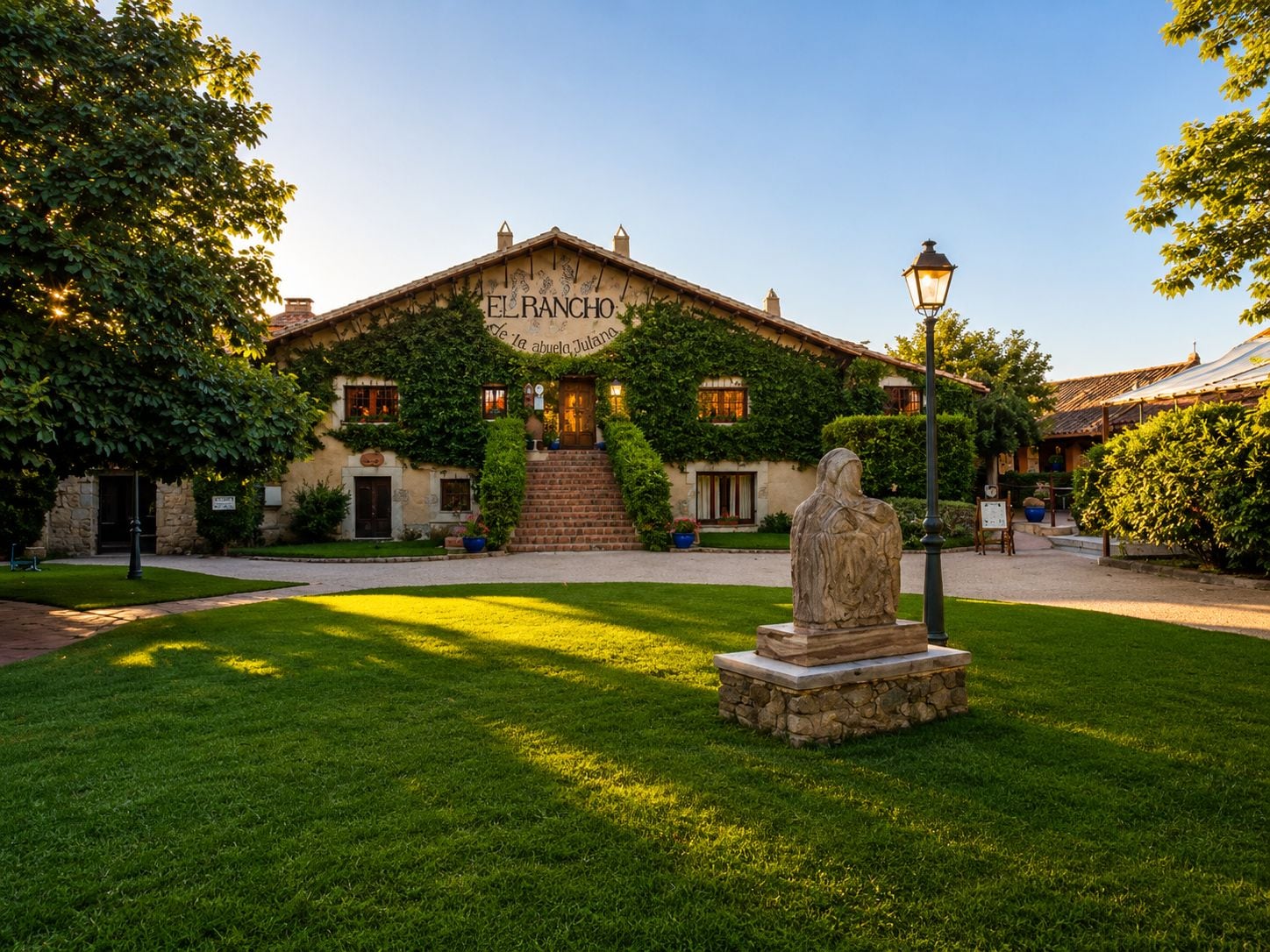 A building named 'El Rancho' with a stone facade, a statue in the foreground, and a well-maintained lawn.
