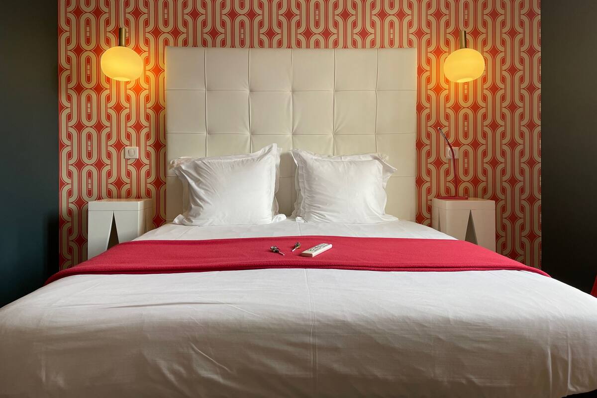 A hotel room with a tufted headboard, white bedding, a red throw blanket, and two bedside lamps.