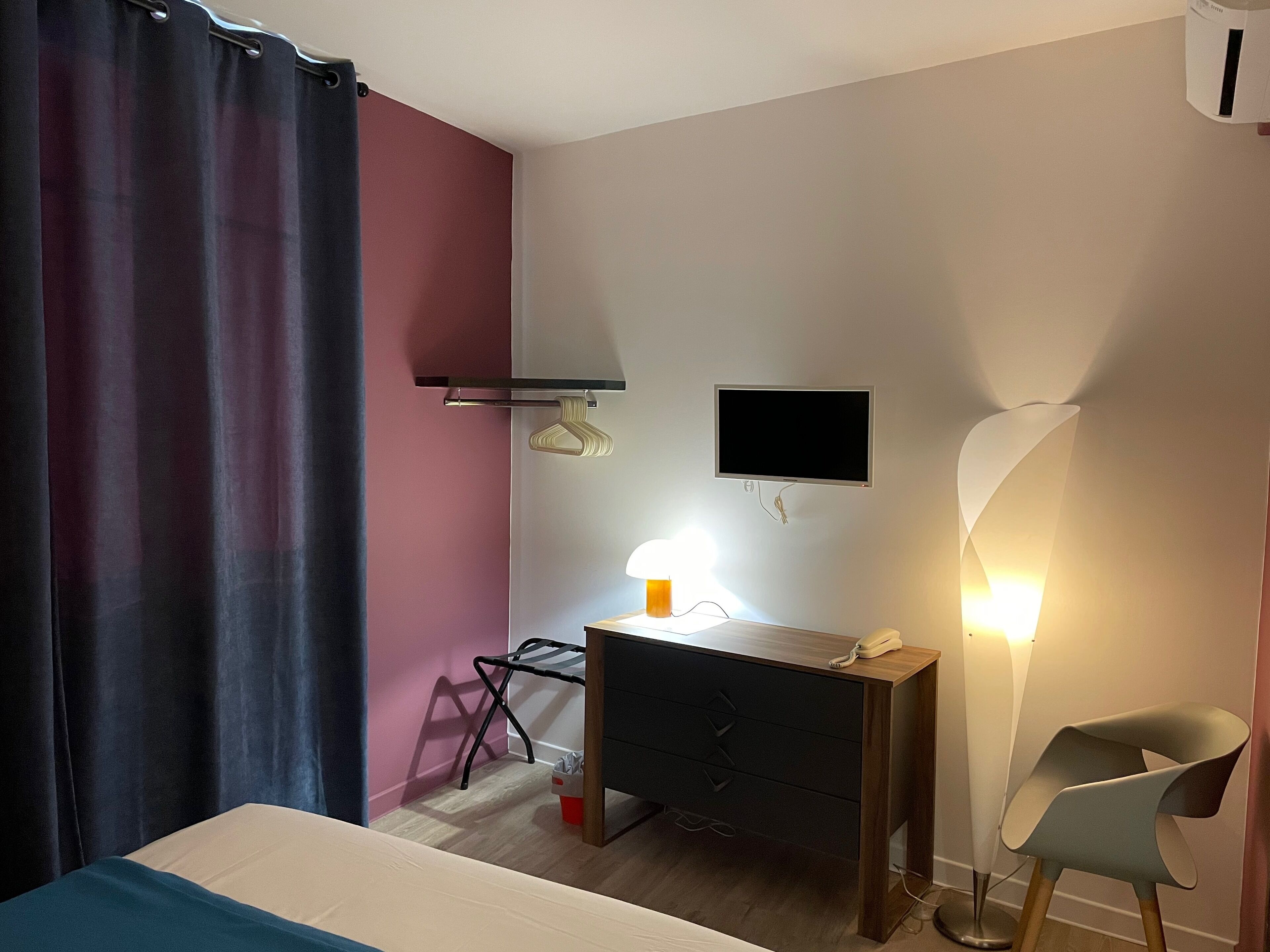 A hotel room with a bed, a desk with a computer, a chair, a TV mounted on the wall, and a bedside lamp.