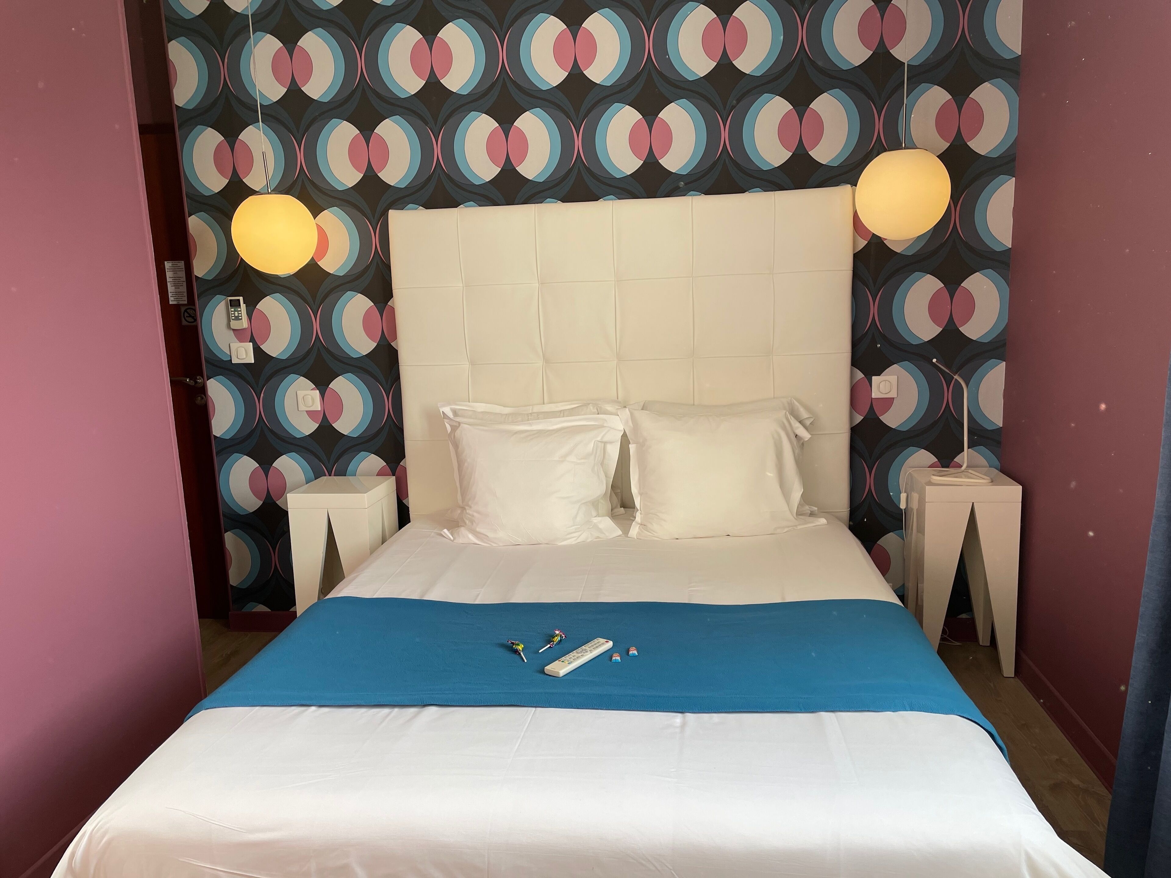 A neatly made bed with white linens, a blue throw blanket, and two pillows. There is a bedside table with a lamp and a phone. The wall behind the bed has a colorful circular pattern.