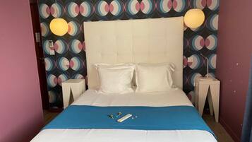 A neatly made bed with white linens, a blue throw blanket, and two pillows. There is a bedside table with a lamp and a phone. The wall behind the bed has a colorful circular pattern.
