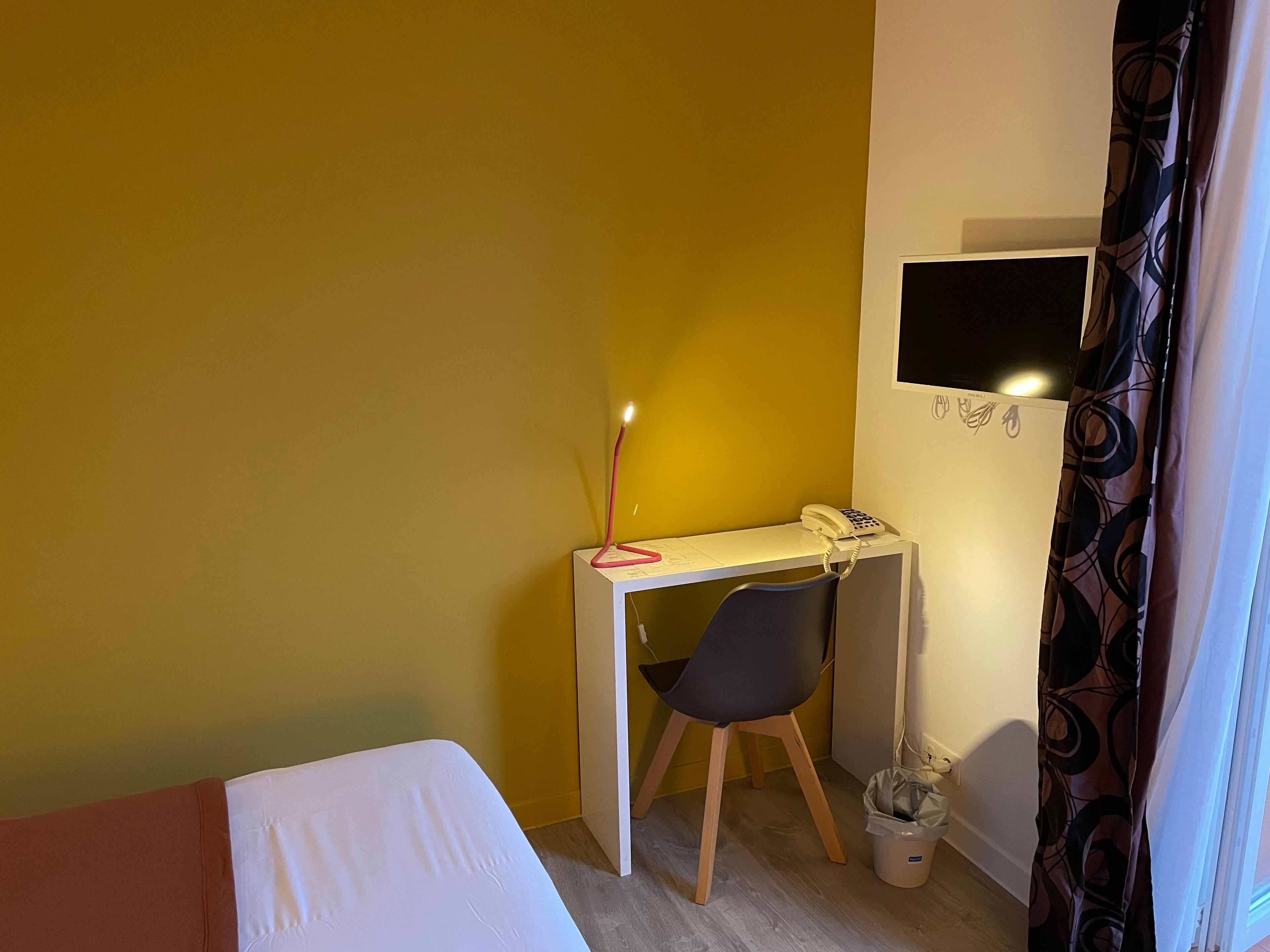 A small room with a bed, a desk with a chair, a telephone, and a TV mounted on the wall.