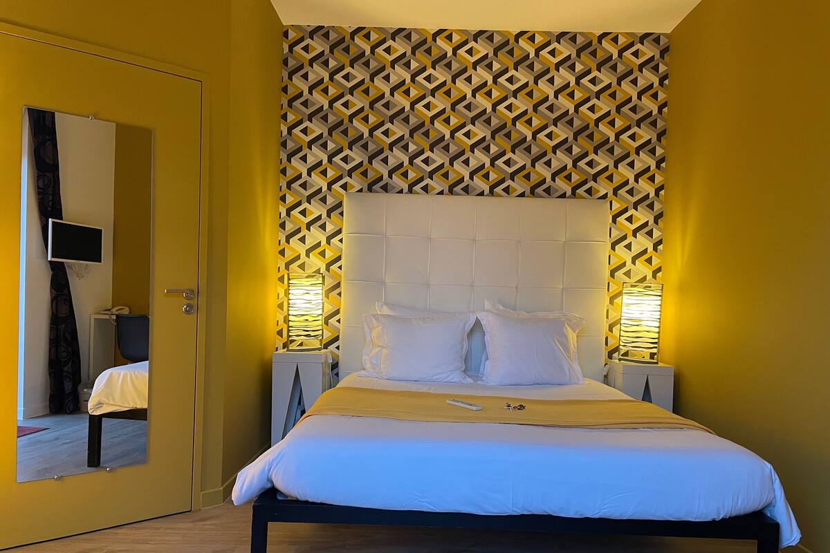 A hotel room with a bed, two bedside lamps, a mirror, a TV, and a patterned wall.