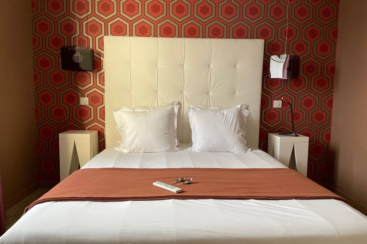 A hotel room with a large bed, two pillows, a remote control, and a nightstand with a lamp.
