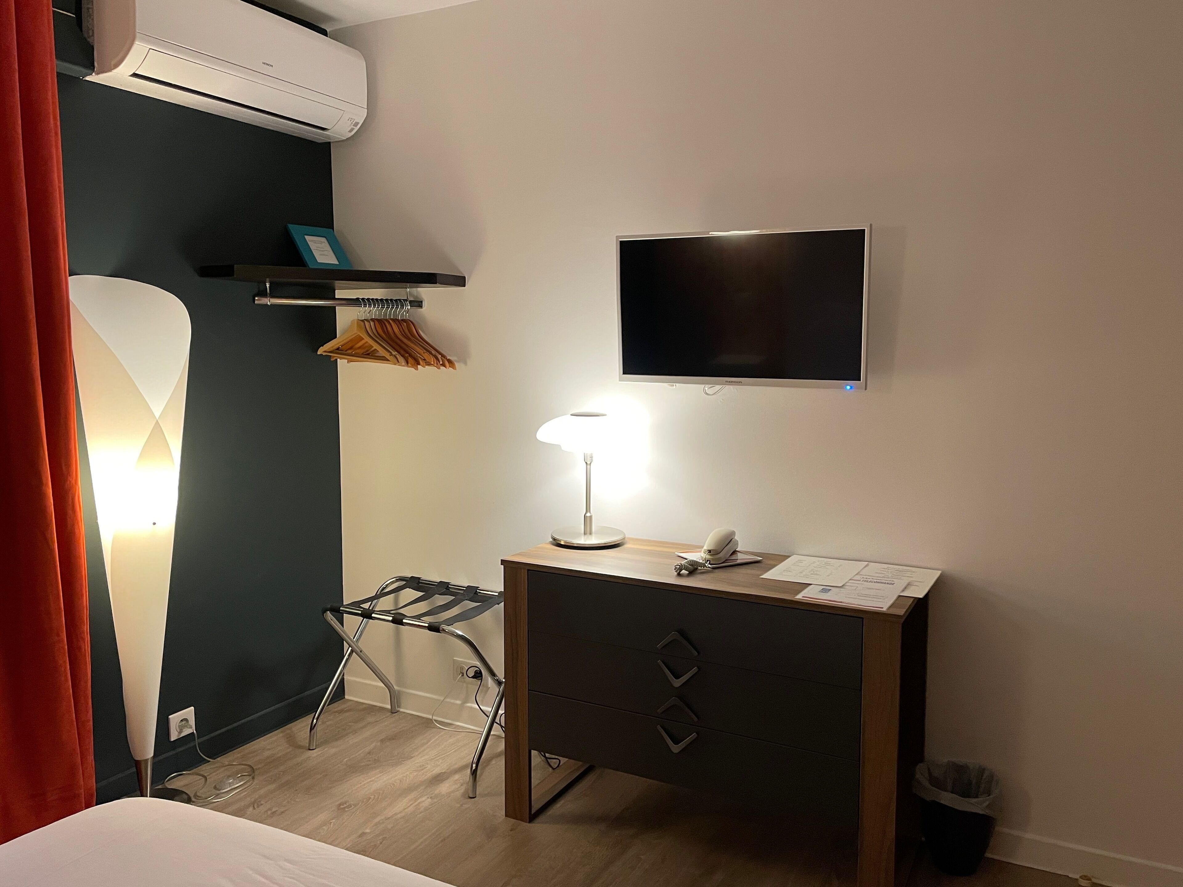 Habitación de hotel con una cama, un escritorio con lámpara, televisión y un pequeño estante con un marco de fotos.