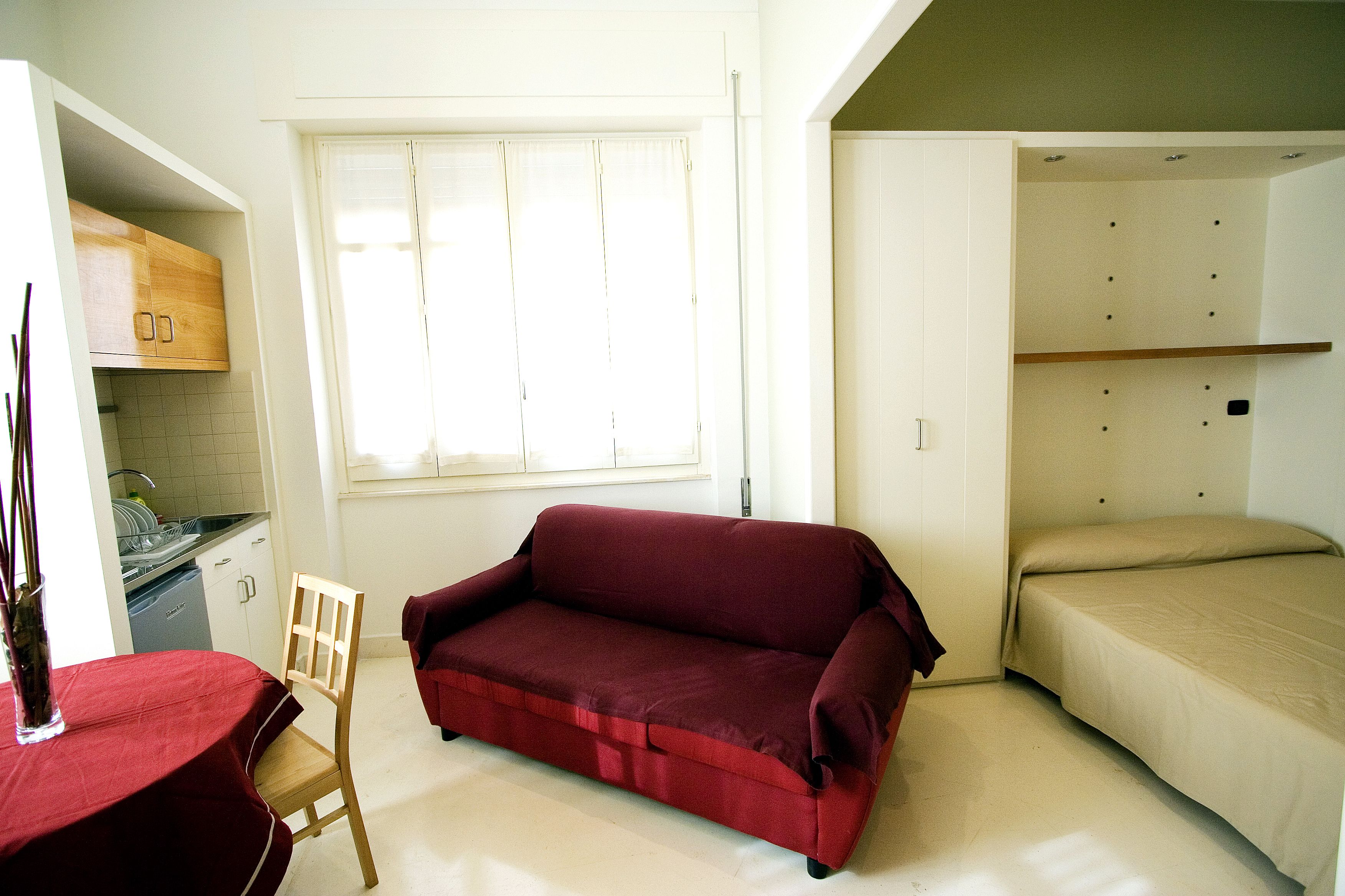A compact room with a kitchenette, a red sofa, a bed, and a window with blinds.