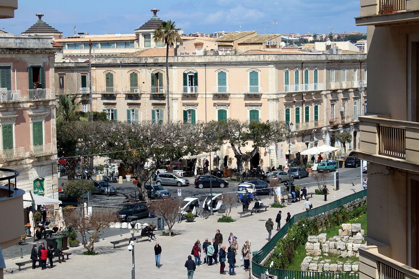 A public square with historic buildings, a central tree, and people walking.