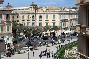 Una plaza pública con edificios históricos, un árbol central y gente caminando.