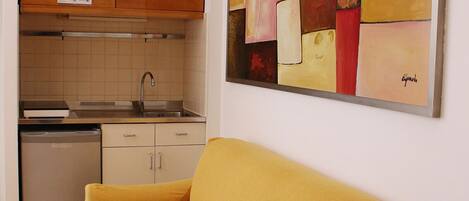 A compact living space with a kitchenette, a yellow sofa, and a colorful abstract painting on the wall.