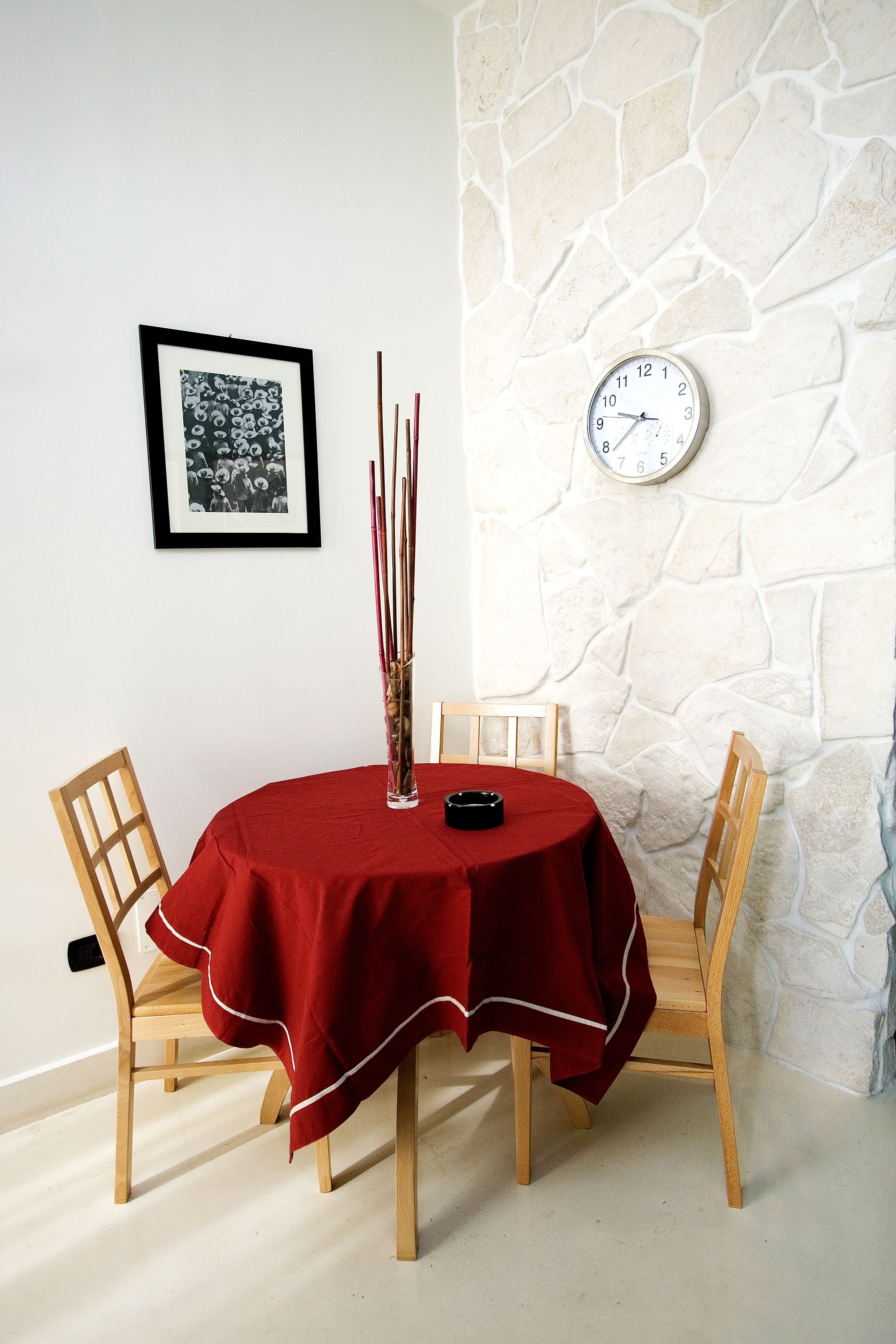 A dining table with a red tablecloth, two wooden chairs, a framed picture, a clock, and a tall vase with sticks.