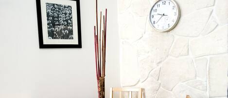 A dining table with a red tablecloth, two wooden chairs, a framed picture, a clock, and a tall vase with sticks.