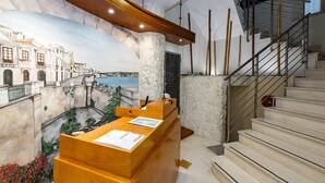 A reception desk with a mural of a coastal town, a stone pillar, and a staircase with metal railings. - Residence Arco Antico (Syracuse)