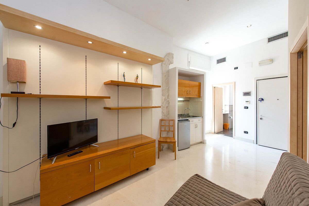 A modern living room with a flat-screen TV on a wooden cabinet, a sofa, and a kitchen area visible through an open door.