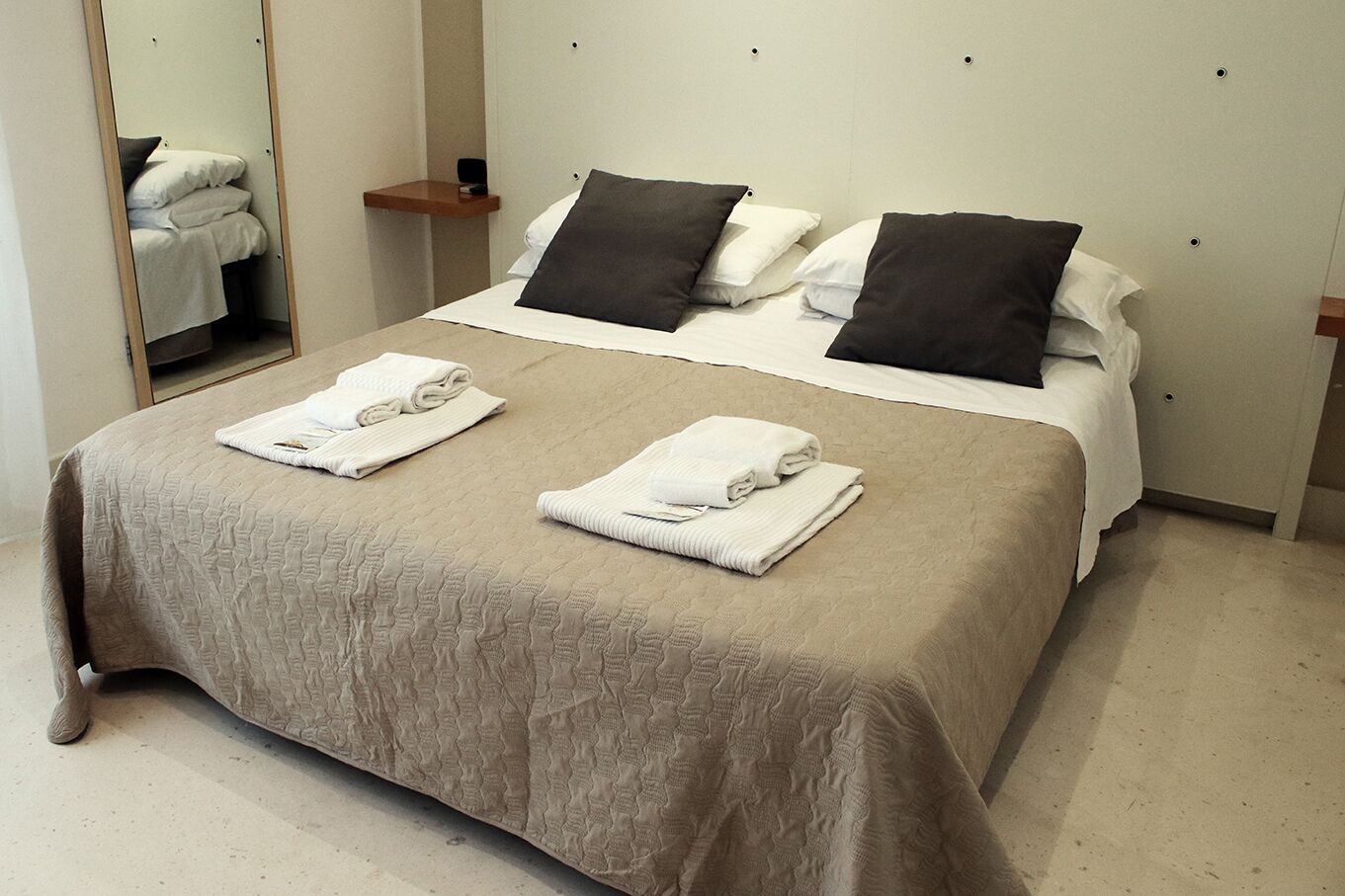Una cama bien hecha con un colcha color beige, dos toallas blancas y dos almohadas negras.