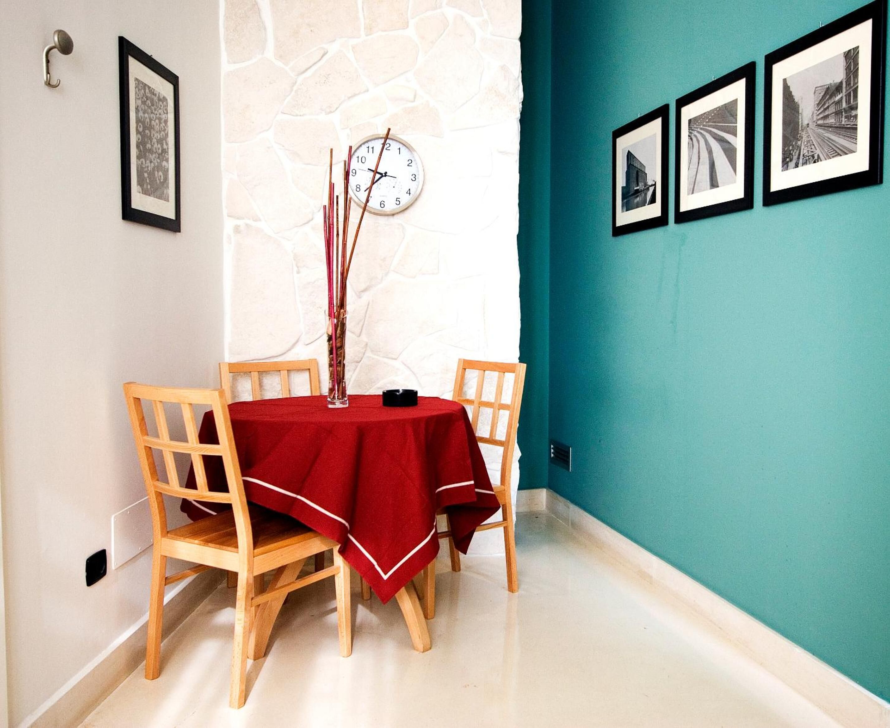 A dining area with a red tablecloth, wooden chairs, a wall clock, and framed pictures on teal walls.