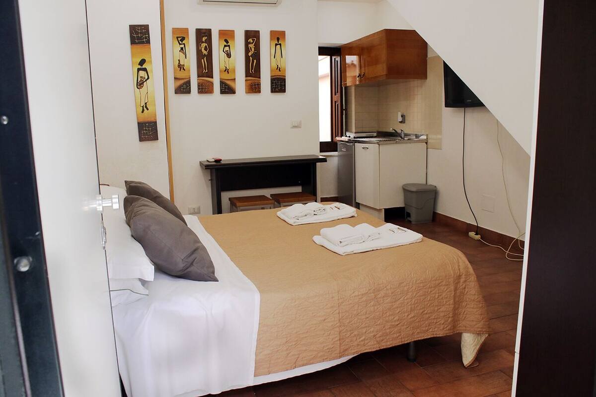 A hotel room with a bed, a desk, a kitchenette, and artwork on the wall.