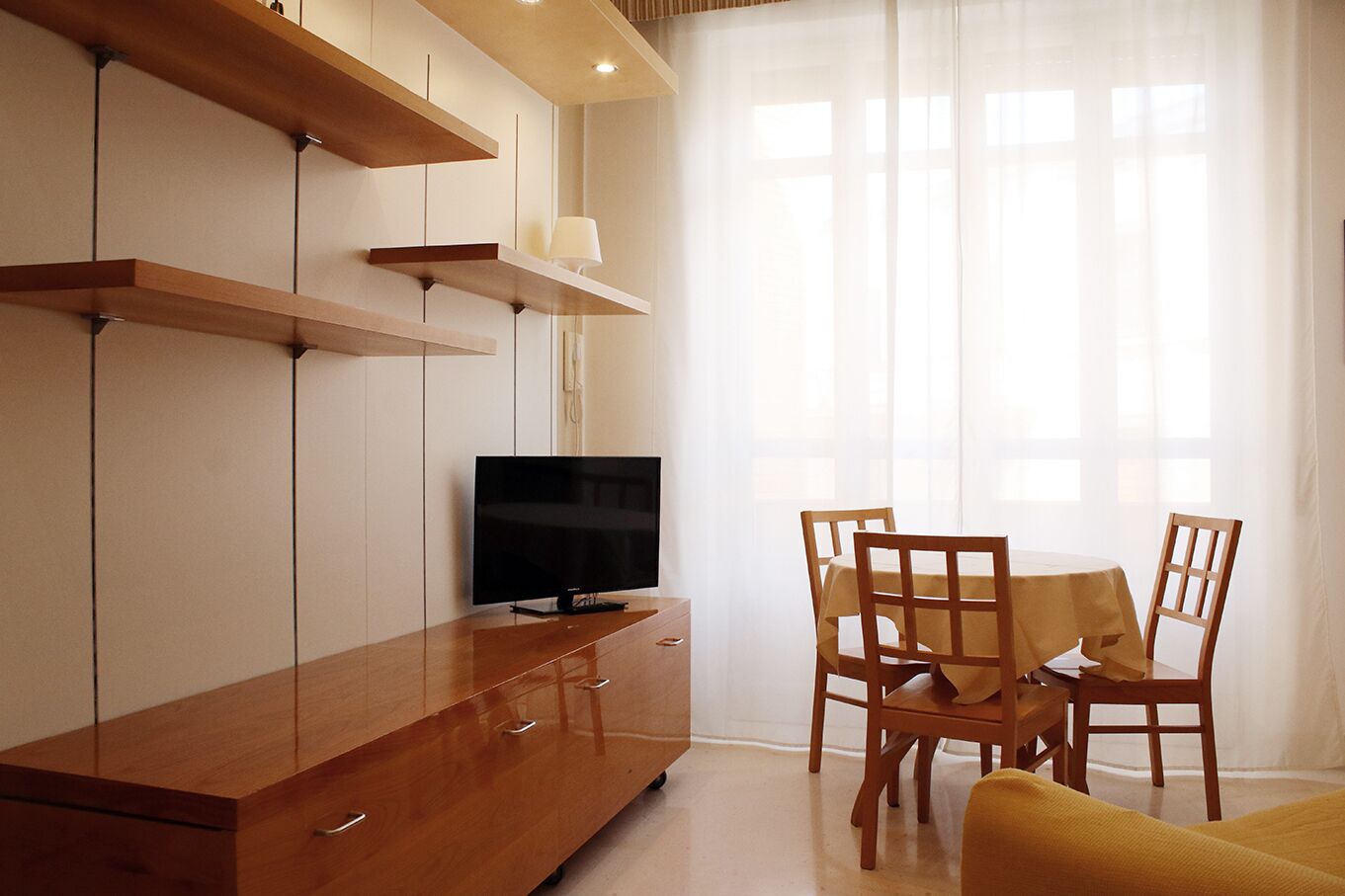 Ein modernes Wohnzimmer mit einem Holz-TV-Möbel, einem Flachbildfernseher, einem Esstisch mit Stühlen und transparenten Vorhängen.