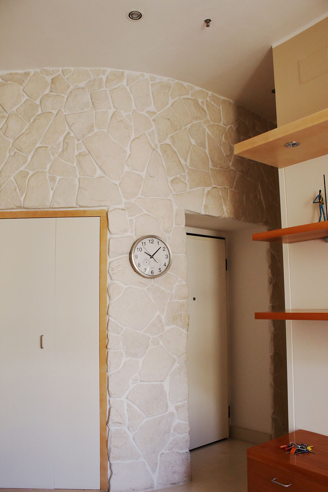 Une pièce avec un mur en pierre, une horloge, une porte et des étagères en bois.
