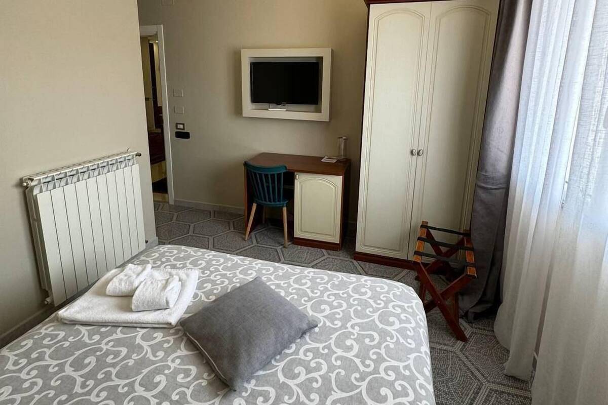 A hotel room with a large bed, a desk with a chair, a wardrobe, a television, and a radiator.