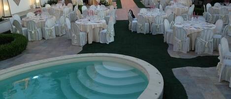 A poolside area with multiple round tables set for dining, surrounded by white chairs.