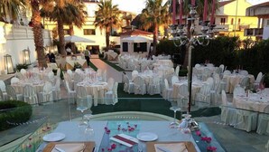An outdoor dining area with round tables set for a formal event, surrounded by palm trees and buildings. - Leon d'Oro (Corleone)