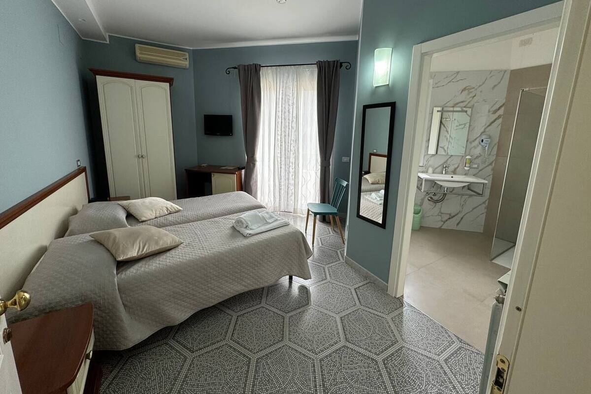 A hotel room with two beds, a bathroom with a marble wall, and a view of the outside through a window.
