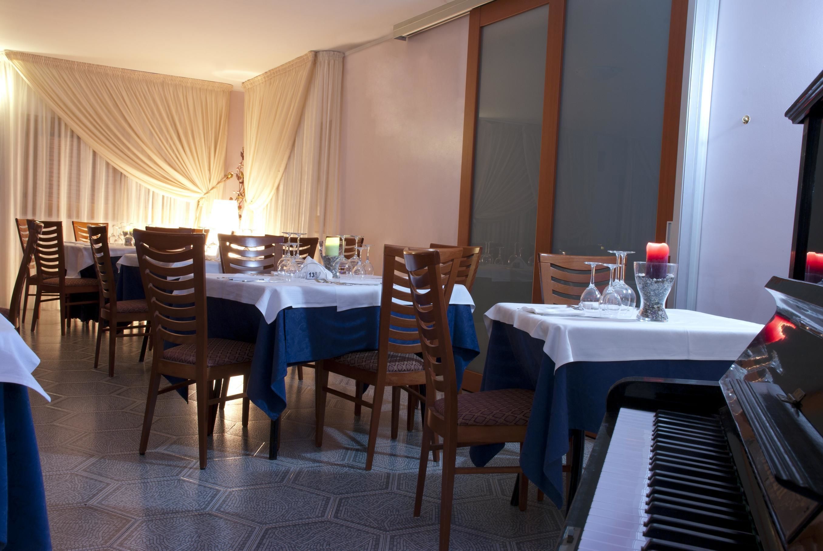 A dining area with tables set for four, wooden chairs, a piano, and a large window with curtains.
