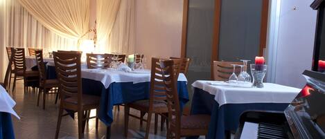A dining area with tables set for four, wooden chairs, a piano, and a large window with curtains.