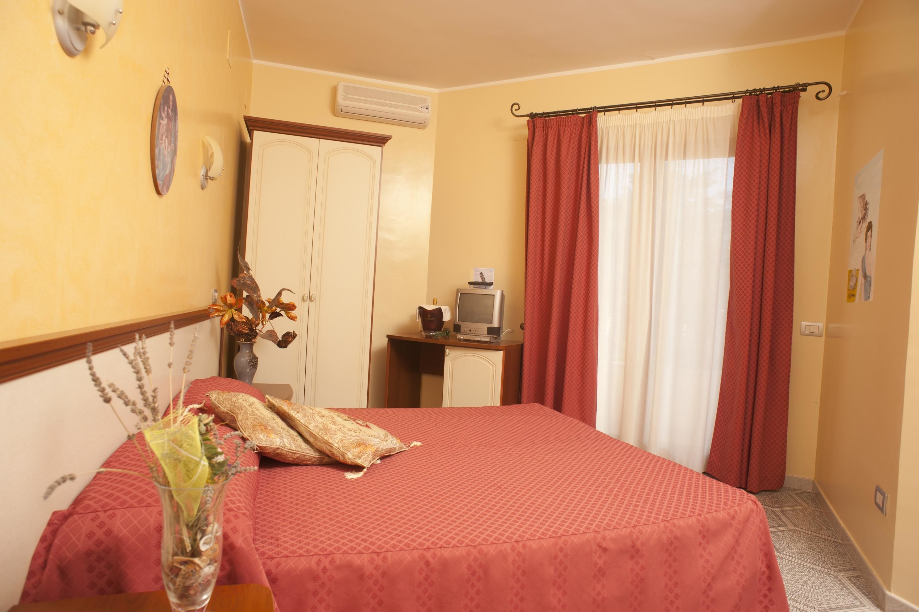 A hotel room with a bed, a desk with a television, a wardrobe, and a window with curtains.