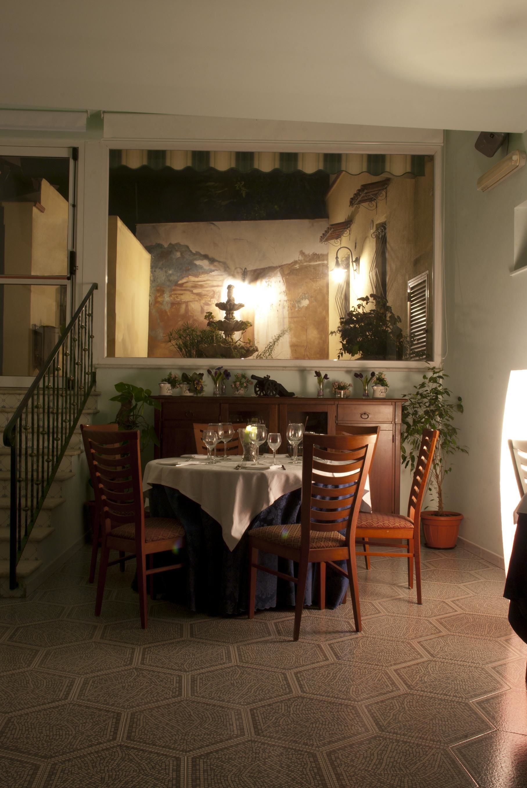 A dining area with a table set for four, surrounded by wooden chairs. A large painting hangs above the table depicting a mountainous landscape with a fountain.