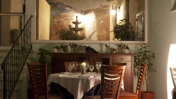 A dining area with a table set for four, surrounded by wooden chairs. A large painting hangs above the table depicting a mountainous landscape with a fountain.