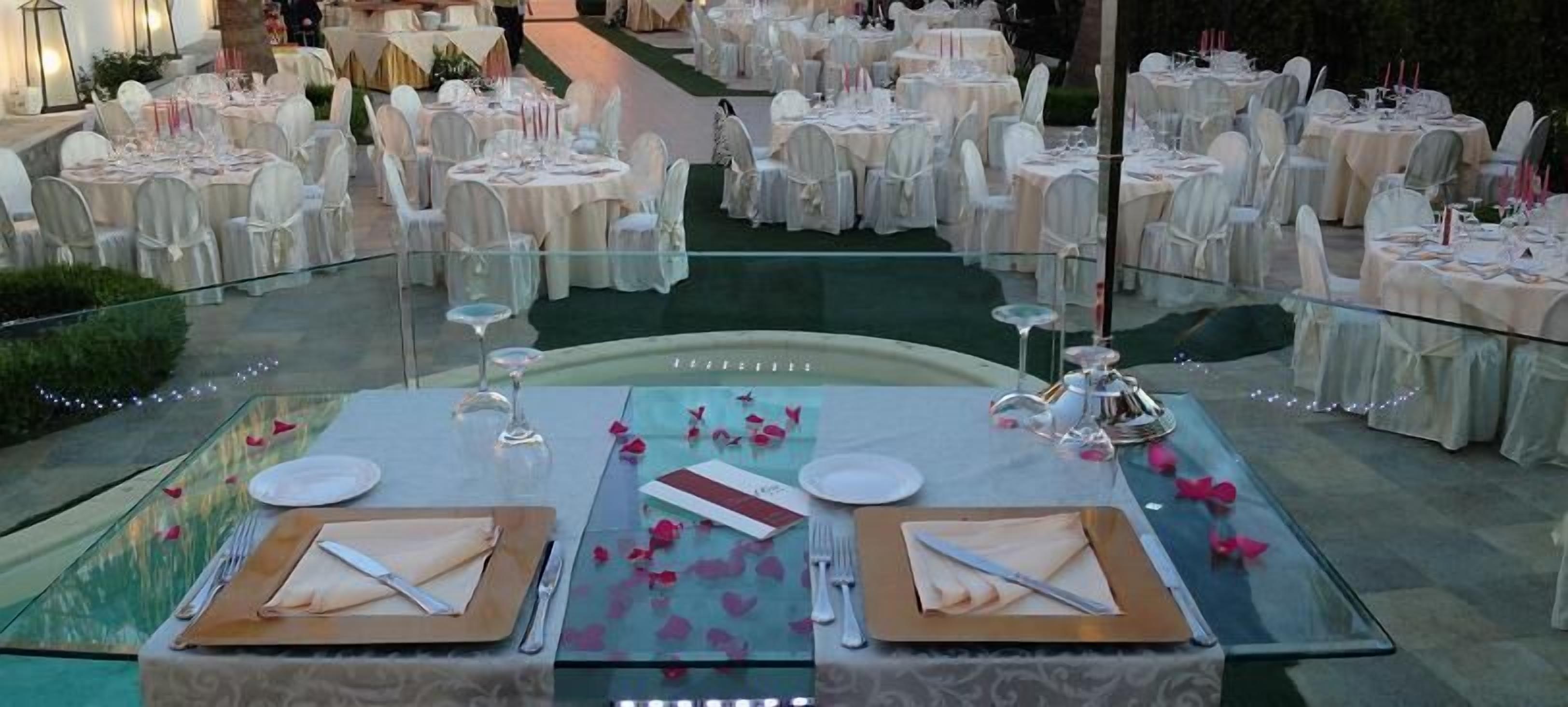 Salão de festas com mesas redondas dispostas para um evento formal, com mesas de vidro e toalhas brancas.