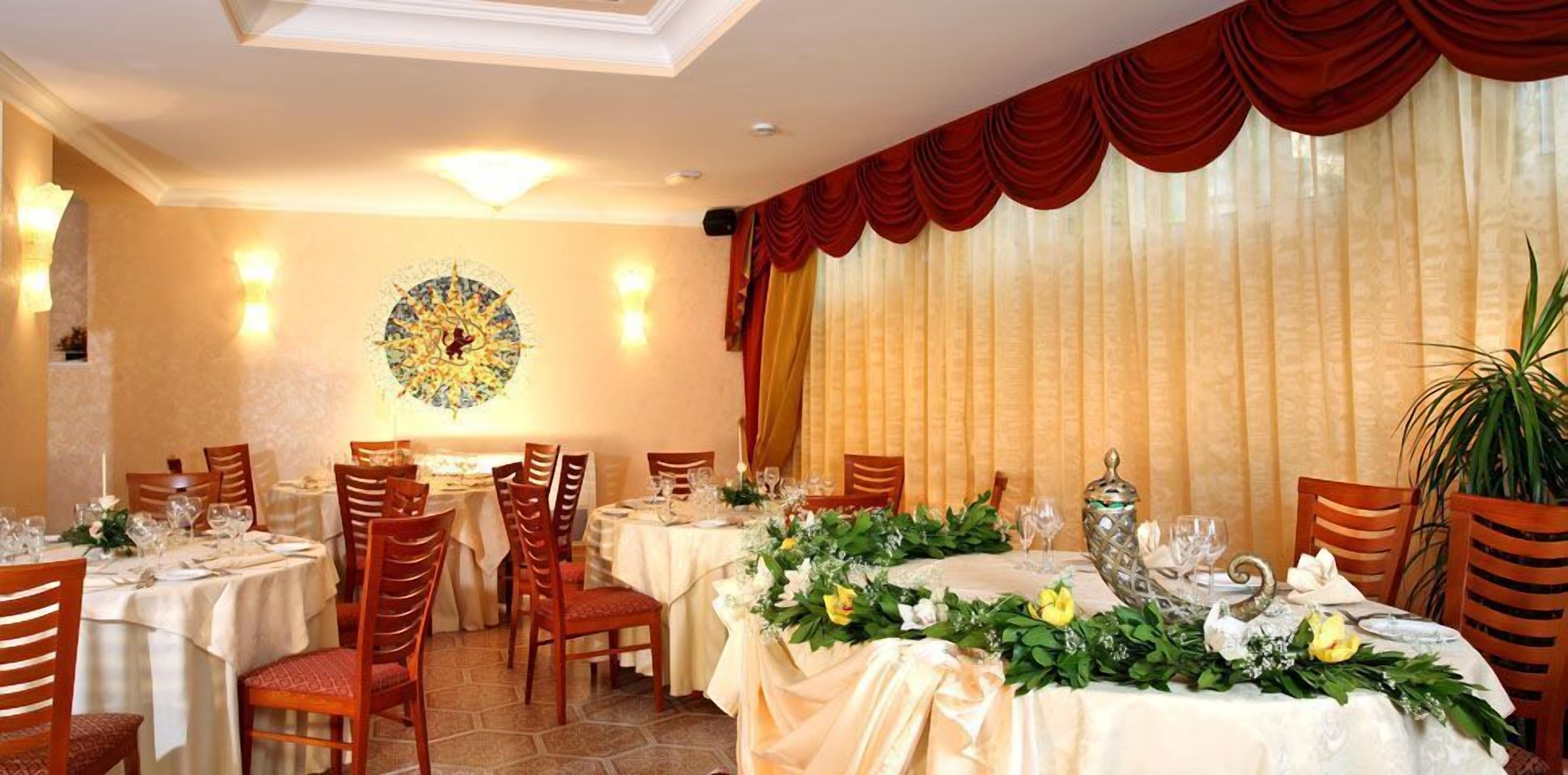 Uma sala de jantar com mesas redondas dispostas para refeição, com toalhas brancas, arranjos florais e cortinas vermelhas.