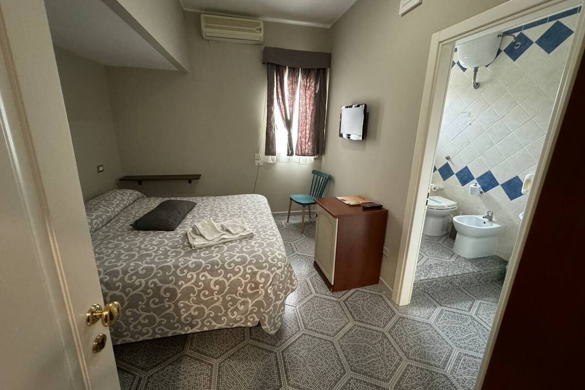 A compact hotel room with a bed, a desk, a chair, a window with curtains, a bathroom with a toilet, bidet, and sink.